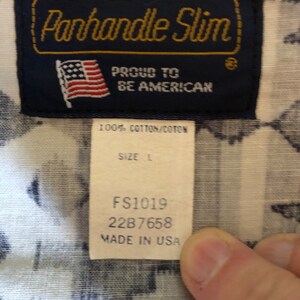 May include: A blue and white label with the text "Panhandle Slim" in gold lettering. The label also includes a small American flag and the text "Proud to be American". Below the label is a white tag with the text "100% Cotton/Coton", "Size L", "FS1019", "2287658", and "Made in USA".