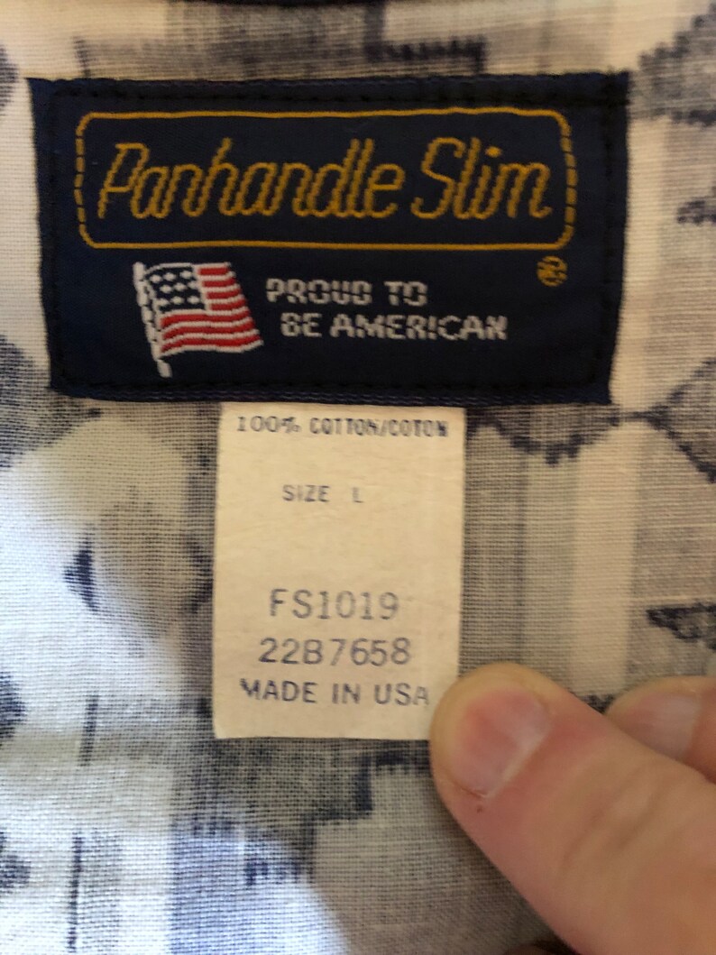 May include: A blue and white label with the text "Panhandle Slim" in gold lettering. The label also includes a small American flag and the text "Proud to be American". Below the label is a white tag with the text "100% Cotton/Coton", "Size L", "FS1019", "2287658", and "Made in USA".