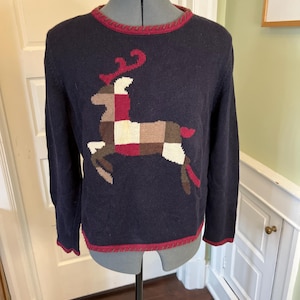 Könnte beinhalten: Ein dunkelblauer Langarmpullover mit einer großen, zentralen Rentiergrafik, die aus einem Patchwork-Muster in Creme-, Beige-, Braun- und Kastanientönen besteht. Der Pullover hat einen Rundhalsausschnitt und einen Saum mit dekorativem kastanienbraunem Zierstich.