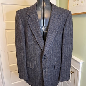May include: A dark gray blazer with a subtle herringbone pattern. The jacket has two front pockets, a notched lapel, and a two-button closure. The blazer is displayed on a mannequin.