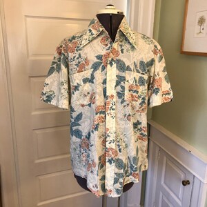 May include: A short-sleeved button-down shirt with a floral print in shades of pink, blue, and green on a white background. The shirt has a collar and a pocket on the left side.