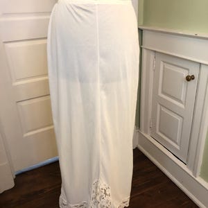 Op de afbeelding: Een lange, witte onderrok met een kanten zoom. De onderrok is gemaakt van een soepele stof en wordt getoond op een paspop. De kanten afwerking voegt een delicate touch toe. De onderrok is in vintage stijl.
