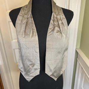 Puede incluir: Un chaleco gris claro con un escote plisado y una parte delantera en forma de V. El chaleco se muestra en un maniquí negro. La tela tiene un brillo sutil y una apariencia texturizada, lo que sugiere seda o un material similar.