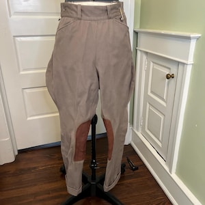 Puede incluir: Pantalones de equitación color topo con diseño de cintura alta, trabillas para cinturón, cierre de cremallera frontal y parches de rodilla en contraste de color marrón tipo ante. El pantalón tiene una silueta acampanada en la cadera.