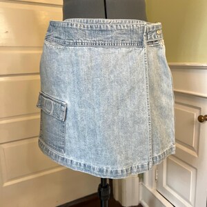 May include: Light blue denim skirt with a cargo pocket on the left side. The skirt is displayed on a mannequin and has a wrap-style design with a button closure. The skirt is made of a light wash denim material.