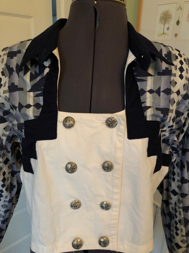 May include: A white button-up shirt with a double row of silver buttons. The shirt has a navy blue collar and cuffs with a geometric pattern in shades of blue and gray.
