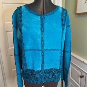 May include: A bright turquoise suede jacket featuring a round neckline, long sleeves, and a front button closure. The garment is constructed with panels of suede joined by dark teal crocheted lace trim, with a wide crocheted lace border at the hem.