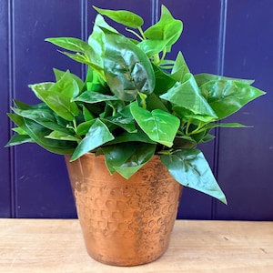May include: A copper-coloured, hammered metal planter filled with vibrant green artificial leaves. The pot has a textured surface and a round shape. The plant is a decorative item for home or office decor.