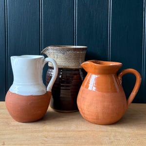 May include: Three ceramic jugs of different sizes and colours. The jug on the left is white and terracotta, the middle one is brown and tan, and the one on the right is orange. All have handles and spouts, set against a dark blue background.