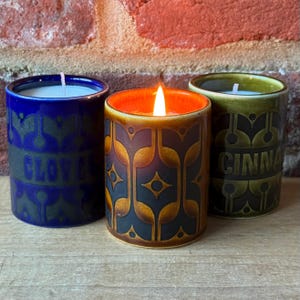 May include: Three ceramic candles in blue, brown, and green. The blue candle has the word "CLOVE" on it, the brown candle is lit, and the green candle has the word "CINNA" on it. Each candle has a decorative pattern.