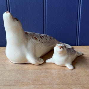 May include: Two ceramic seal ornaments. The larger seal is lying down with its head raised, and the smaller seal is lying down. Both seals are off-white with brown speckled markings.
