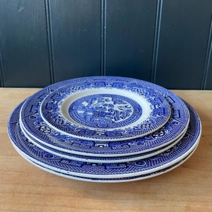 May include: A stack of three blue and white ceramic plates with a classic Willow pattern. The plates are arranged in a nested configuration, with the smallest plate on top. The plates are on a wooden surface.