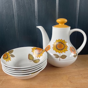 May include: A white ceramic teapot with a yellow lid and a matching stack of four white bowls. Both the teapot and the bowls feature a decorative pattern of yellow and orange sunflowers with green leaves painted on the exterior surfaces.