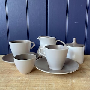 May include: A collection of neutral-toned ceramic tableware. The set comprises two teacups, two saucers, a small bowl, a creamer jug, and a lidded sugar bowl. The inside of the cups and bowl are a darker grey colour.