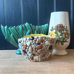 May include: A selection of vintage ceramic vases and bowls. The focal point is a small bowl with a pebble design and a yellow rim. A tall, cream-coloured vase with floral patterns and a green, leaf-shaped bowl complete the arrangement.