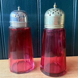 May include: A pair of vintage salt shakers. They feature ruby red glass bodies and silver-coloured metal lids. The glass has a faceted design, and the lids are perforated. The shakers are displayed on a wooden surface.