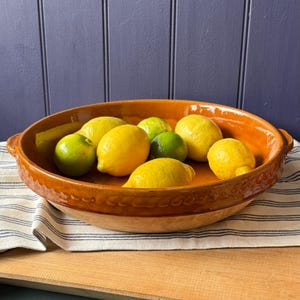 May include: A terracotta serving bowl filled with lemons and limes. The bowl has a decorative rim and handles. The lemons are yellow, and the limes are green. The bowl sits on a striped cloth. The bowl is approximately 30 cm in diameter.