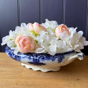 May include: A white and blue ceramic oval serving dish with ornate dark blue scrollwork patterns along the rim, filled with a dense arrangement of artificial white hydrangea blossoms and several soft peach-coloured rose buds.