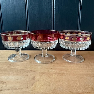 May include: Three vintage clear glass dessert bowls with ruby red rims and gold floral accents. The bowls have a clear, textured base and a stem. The bowls are displayed on a wooden surface.