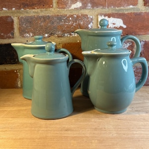 May include: Four teal ceramic pitchers with lids. Each pitcher has a handle and a spout. The pitchers are arranged on a wooden surface in front of a brick wall.