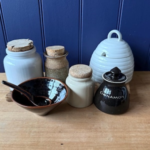 May include: A selection of ceramic kitchen items, including jars with cork lids, a small bowl with a spoon, and a white honey pot. The black jar is labelled "CINNAMON". The items are arranged on a wooden surface.