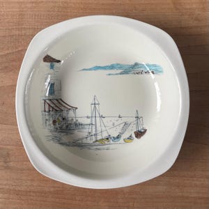 May include: A white ceramic bowl with a coastal scene illustration. The design features a harbour with boats, a pier, and a building with a striped awning. The bowl has a slightly square shape with rounded corners.