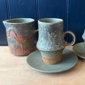 May include: Handmade ceramic mug and creamer set. The mug has a speckled green and brown glaze with a handle and sits on a matching saucer. The creamer has a unique design with a handle and a textured, earthy appearance. The set is perfect for coffee or tea.