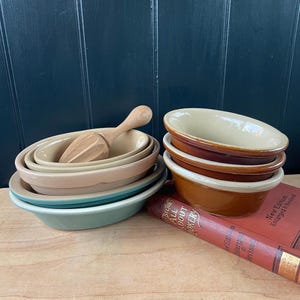 May include: A selection of vintage kitchen items, featuring oval ceramic bowls in beige, brown, and teal, a wooden citrus reamer, and a stack of cookbooks. The cookbooks are red with gold lettering. The bowls are stacked together.