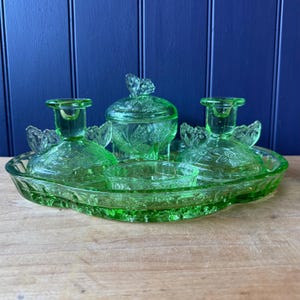 May include: A set of light green pressed glass vanity items on a matching oval tray. The collection includes two candle holders, a lidded jar, and a small open bowl, all featuring embossed butterfly and floral motifs throughout the glass surface.