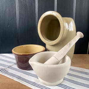May include: A selection of antique kitchen items, featuring a mortar and pestle, a small brown and tan bowl, and a large tan ceramic container. The items are displayed on a striped cloth.