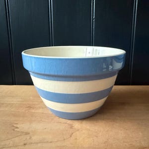May include: A blue and white striped ceramic mixing bowl. The bowl has a wide, flared shape with a light blue rim and alternating blue and white horizontal stripes. The bowl sits on a wooden surface.