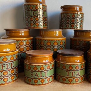 May include: A collection of ceramic storage canisters featuring a brown and green geometric pattern and wooden lids. Some canisters are labelled with the words SUGAR, COFFEE, SALT, and TEA in embossed lettering. The set includes various sizes.