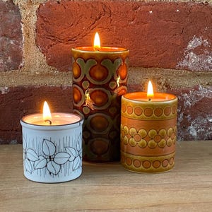 May include: Three lit ceramic candles with warm orange flames. The candles are cylindrical, with decorative patterns in shades of brown, orange, and white. One candle has a floral design, another has a geometric pattern, and the third has a leaf design.