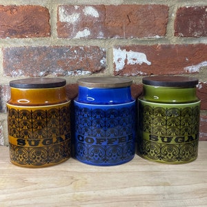May include: Three ceramic canisters with black and white patterned designs. The canisters are labelled "Sugar", "Coffee", and "Sugar". The canisters are brown, blue, and green.