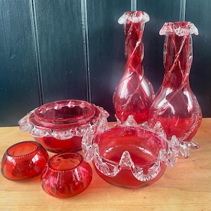 May include: A set of ruby red glass vases and bowls. Two tall vases feature a twisted design, and the bowls have ruffled edges. The glass pieces are displayed on a wooden surface, highlighting their intricate details and vibrant colour.
