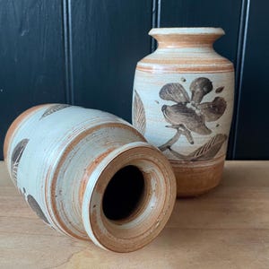 May include: Two ceramic vases with a brown and white floral design. The vases are a light brown colour with a white glaze and have a brown floral design painted on them.