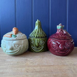 May include: Three ceramic lidded storage jars shaped like vegetables with anthropomorphic faces. The left jar is light green with a carrot topper, the centre jar is dark green labelled CUCUMBER, and the right jar is deep red labelled BEETROOT.