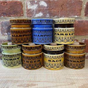 May include: A selection of ceramic spice jars in shades of brown, blue, and yellow. Each jar is labelled with a spice name in black text and features a decorative floral design. The jars have wooden lids.