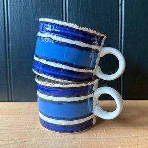 May include: Two stacked ceramic mugs with a blue and white striped design. The mugs have a rounded shape with a white handle. The interior of the mugs is a lighter shade of blue. The rims of the mugs have a brown, textured edge.