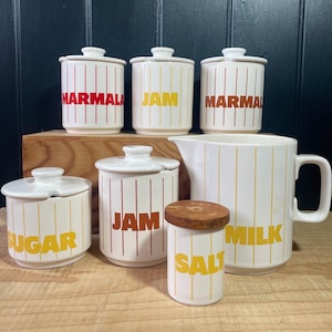 May include: A set of vintage kitchen canisters with yellow and red striped designs. The canisters are labelled "JAM", "MARMALADE", "SUGAR", and "MILK". There is also a smaller canister labelled "SALT" with a wooden lid and a white jug.