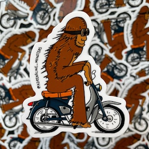 Könnte beinhalten: Ein Aufkleber mit einem braunen Bigfoot-Charakter mit Brille und Helm, der ein blaues und graues Motorrad fährt. Der Aufkleber trägt den Text "@adrenaline_monsters". Der Hintergrund ist mit ähnlichen Aufklebern gefüllt.