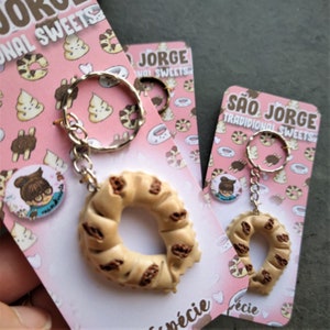 May include: Keychains featuring a miniature, beige, ring-shaped pastry with dark brown accents, attached to a silver keyring. The keychains are displayed on pink cards with the text "SÃO JORGE TRADITIONAL SWEETS" and illustrations of pastries.