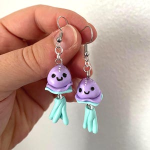 May include: A pair of handmade earrings featuring a whimsical jellyfish design. Each earring has a lavender-colored head with a smiling face, a light blue frill, and matching tentacles. The earrings are suspended from silver-colored hooks.
