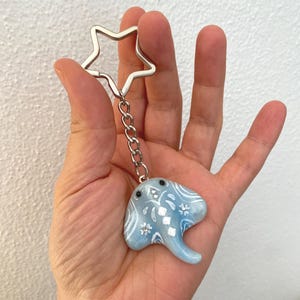 May include: A light blue stingray keychain with white floral and geometric designs. The keychain has a silver chain and a star-shaped keyring. The stingray has black dot eyes and a curved tail.