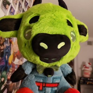Pode incluir: Um brinquedo de pelúcia de vaca com uma cabeça verde brilhante, chifres pretos e um focinho preto. A vaca usa uma roupa azul clara com um logotipo vermelho e branco. Tem pés vermelhos e orelhas pretas. O brinquedo tem uma gravata borboleta e olhos grandes e claros.