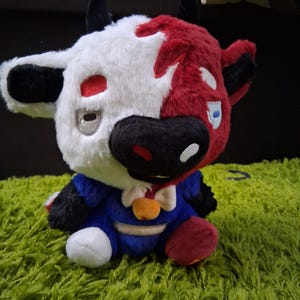May include: A plush toy cow with a black and white face and a red and white body. The cow is wearing a blue suit with a white bow tie and a gold button. The cow is sitting on a green carpet.