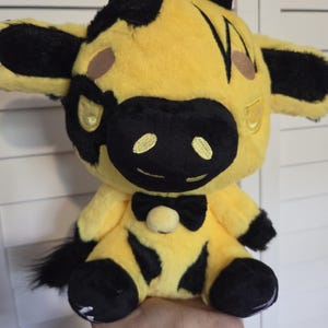 May include: A plush cow toy with a yellow body and black spots, ears, and muzzle. The cow has a black bow tie and a small tail. The toy is held in a person's hand.