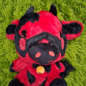 May include: A red and black plush toy cow with horns, a black bow tie, and a yellow bell. The cow is sitting on a green shag carpet.