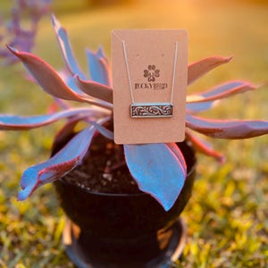 Puede incluir: Un collar de plata con un colgante de barra decorativo y adornado. El colgante tiene un diseño floral y el texto "LUCKY BIRD" está grabado en la barra. El collar se muestra en una tarjeta marrón con un trébol verde.