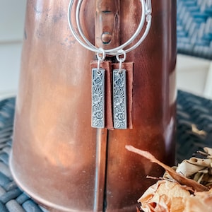 May include: Silver hoop earrings with a floral design dangle from the hoops. The dangle is a silver bar with a floral design and a brown leather backing.